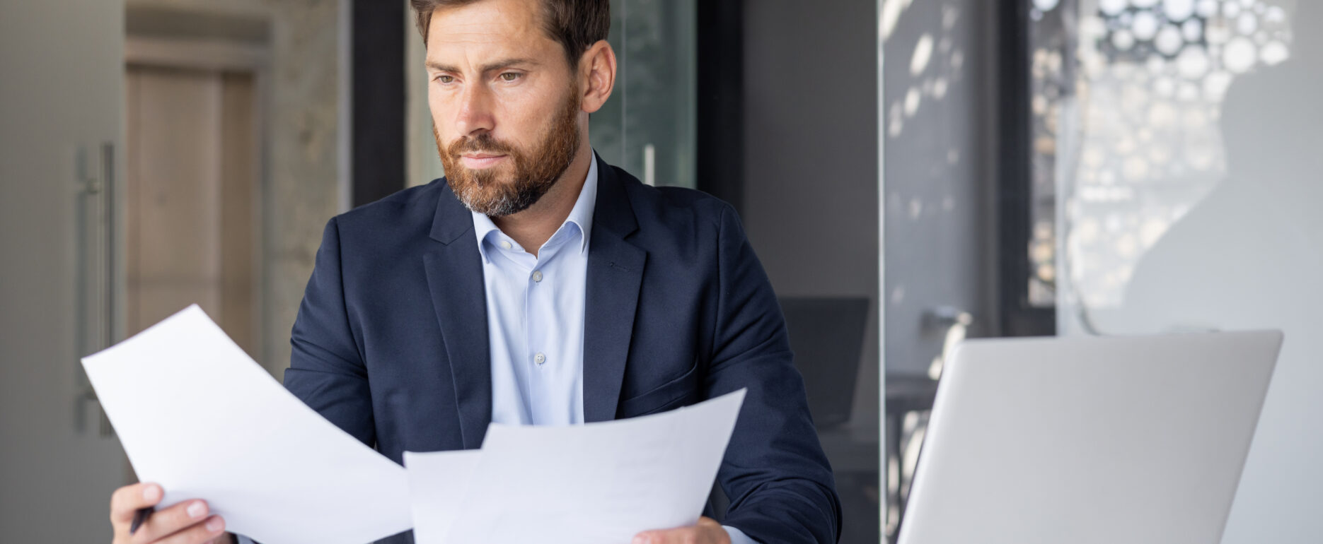 Serious and concentrated young man businessman sitting in the office at the desk with a laptop in a suit and working with documents and a project.